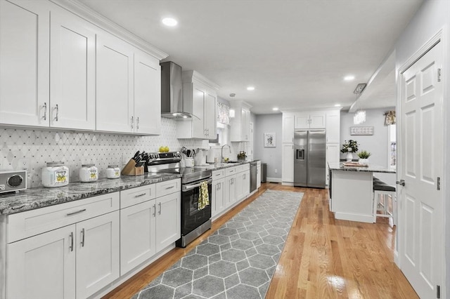 kitchen featuring a breakfast bar, a sink, stainless steel appliances, white cabinets, and wall chimney range hood