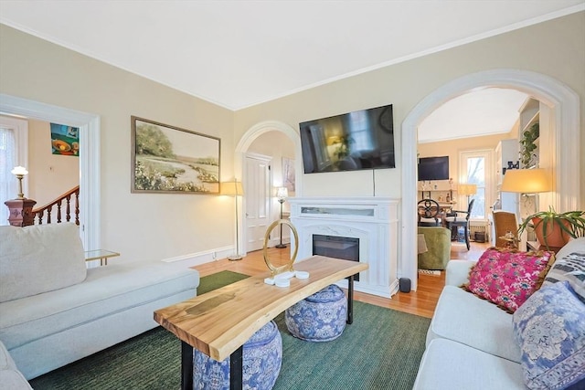 living area with ornamental molding, wood finished floors, a glass covered fireplace, arched walkways, and stairs