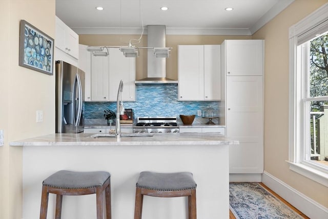kitchen featuring stainless steel fridge with ice dispenser, stove, wall chimney exhaust hood, a breakfast bar, and recessed lighting