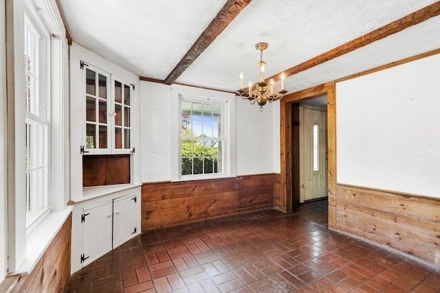 unfurnished room with a chandelier, wainscoting, wooden walls, beam ceiling, and brick patterned floors