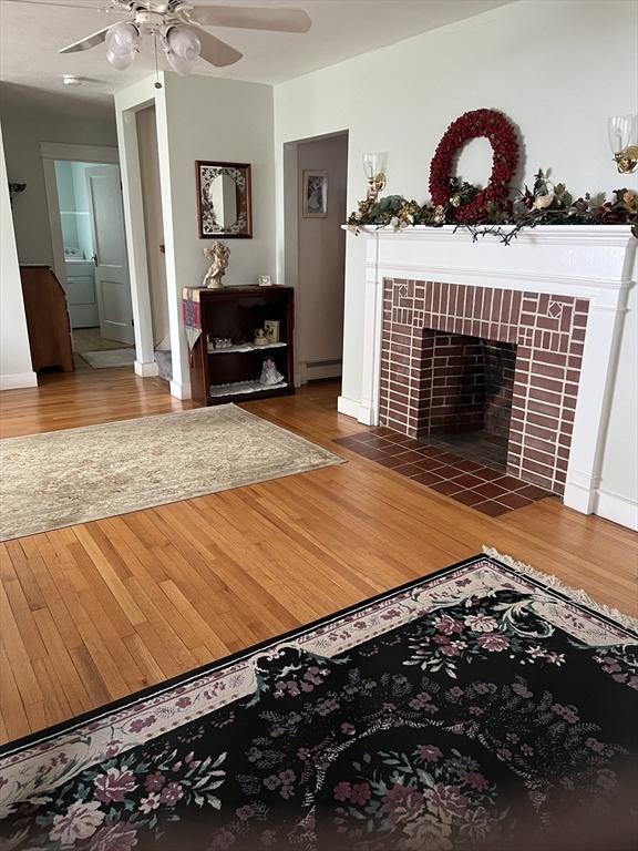 living area with a ceiling fan, baseboard heating, washer / clothes dryer, a tiled fireplace, and wood finished floors