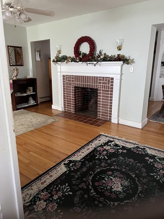 living area with wood finished floors, a baseboard heating unit, a fireplace, a ceiling fan, and baseboards