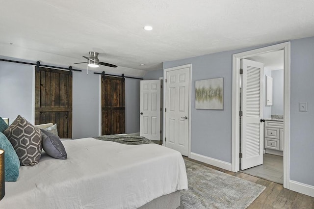 bedroom featuring light wood-style flooring, a barn door, ceiling fan, ensuite bath, and baseboards