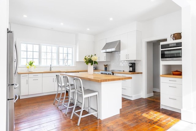 kitchen with white cabinetry, wooden counters, appliances with stainless steel finishes, wall chimney exhaust hood, and tasteful backsplash