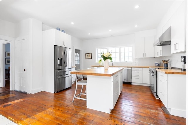 kitchen with stainless steel appliances, wood counters, a kitchen breakfast bar, wall chimney range hood, and backsplash