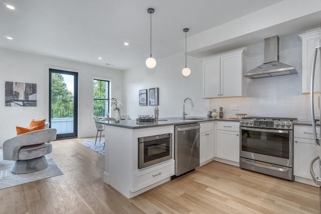 kitchen with a peninsula, light wood-style flooring, a sink, stainless steel appliances, and wall chimney exhaust hood