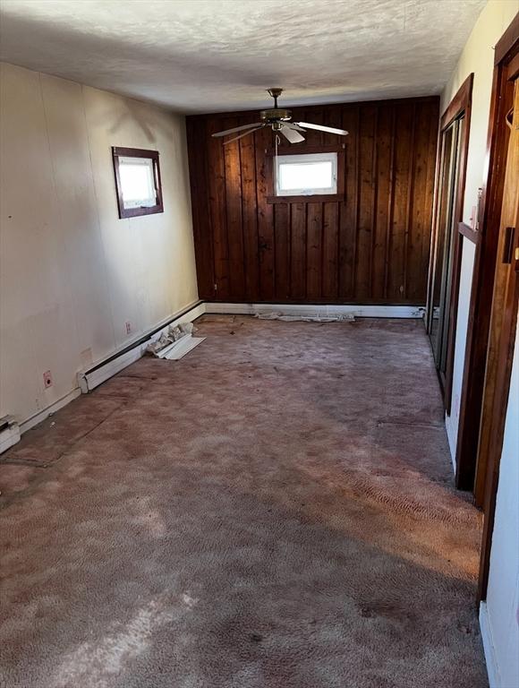 unfurnished room featuring a textured ceiling, wood walls, baseboard heating, and dark colored carpet