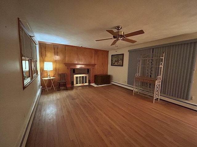 unfurnished living room featuring hardwood / wood-style flooring, ceiling fan, a baseboard heating unit, a brick fireplace, and wood walls