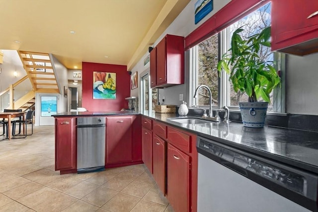 kitchen with light tile patterned floors, dark countertops, stainless steel dishwasher, a sink, and a peninsula