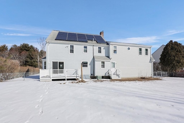 snow covered back of property featuring central AC unit, solar panels, a chimney, fence, and a wooden deck