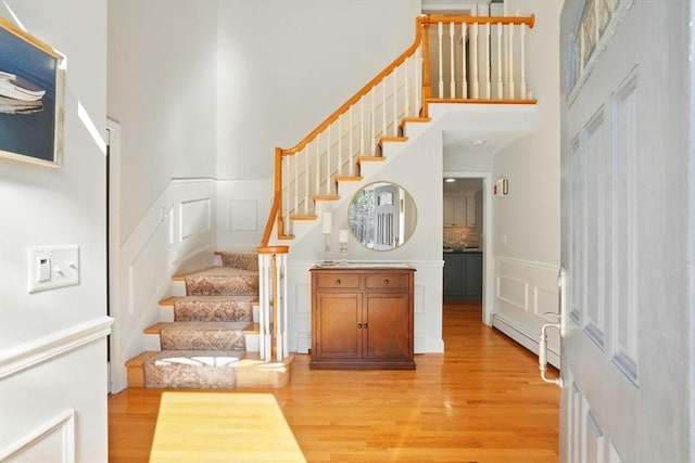 staircase featuring a towering ceiling, a baseboard radiator, wood finished floors, and a decorative wall