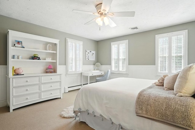 carpeted bedroom with a ceiling fan, a textured ceiling, visible vents, wainscoting, and a baseboard radiator