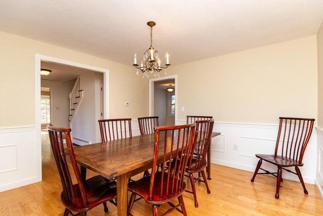 dining area featuring a notable chandelier, wainscoting, baseboard heating, light wood-style floors, and stairway