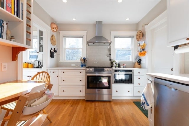 kitchen with stainless steel appliances, white cabinets, light wood-style floors, and wall chimney range hood