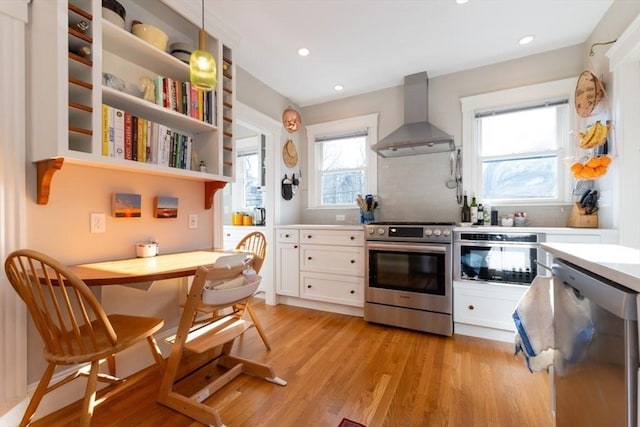 kitchen with appliances with stainless steel finishes, white cabinets, light wood-style floors, and wall chimney exhaust hood