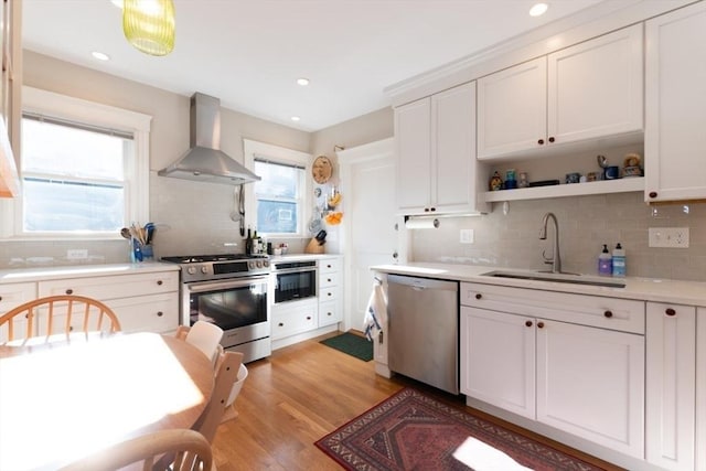 kitchen featuring stainless steel appliances, a sink, light countertops, light wood-type flooring, and wall chimney exhaust hood