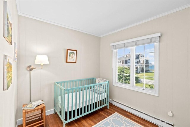 bedroom featuring a baseboard radiator, wood finished floors, crown molding, a nursery area, and baseboards