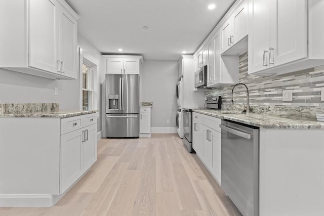 kitchen with backsplash, appliances with stainless steel finishes, stacked washing maching and dryer, white cabinets, and light wood-style floors