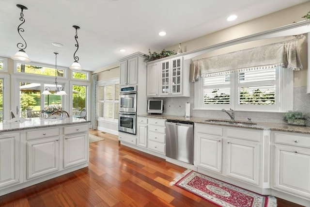 kitchen with a sink, appliances with stainless steel finishes, glass insert cabinets, wood finished floors, and decorative light fixtures