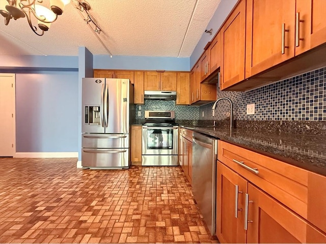 kitchen with appliances with stainless steel finishes, under cabinet range hood, a sink, backsplash, and brown cabinetry