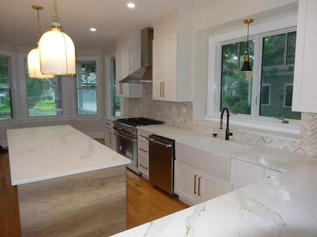 kitchen with light stone counters, white cabinetry, hanging light fixtures, stainless steel appliances, and wall chimney range hood