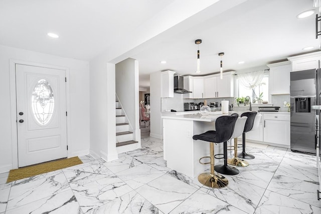 kitchen featuring stainless steel fridge, tasteful backsplash, marble finish floor, wall chimney range hood, and a kitchen bar