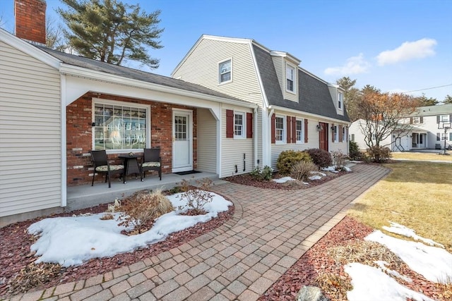 exterior space featuring brick siding, roof with shingles, covered porch, a lawn, and a gambrel roof