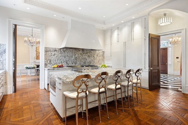 kitchen featuring premium range hood, a kitchen island with sink, a chandelier, and white cabinets