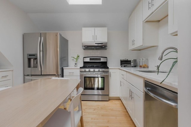 kitchen with a skylight, a sink, under cabinet range hood, appliances with stainless steel finishes, and backsplash