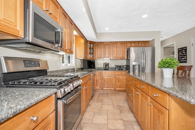 kitchen with appliances with stainless steel finishes, a sink, stone tile floors, dark stone countertops, and brown cabinetry