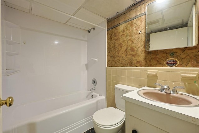 bathroom with toilet, a wainscoted wall,  shower combination, tile walls, and wallpapered walls