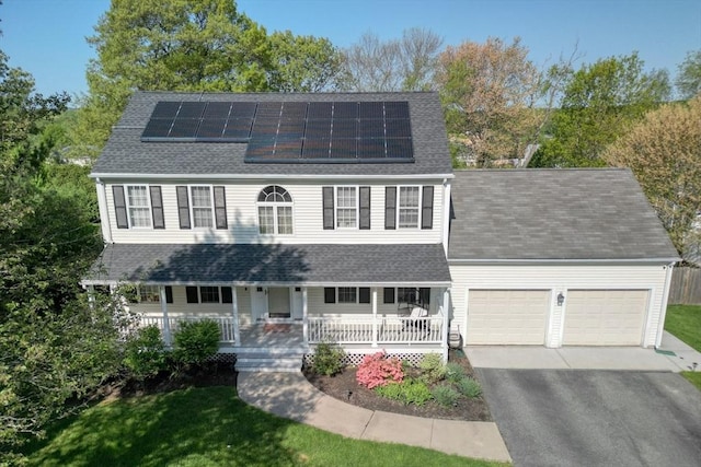 colonial-style house with a garage, asphalt driveway, covered porch, roof mounted solar panels, and roof with shingles