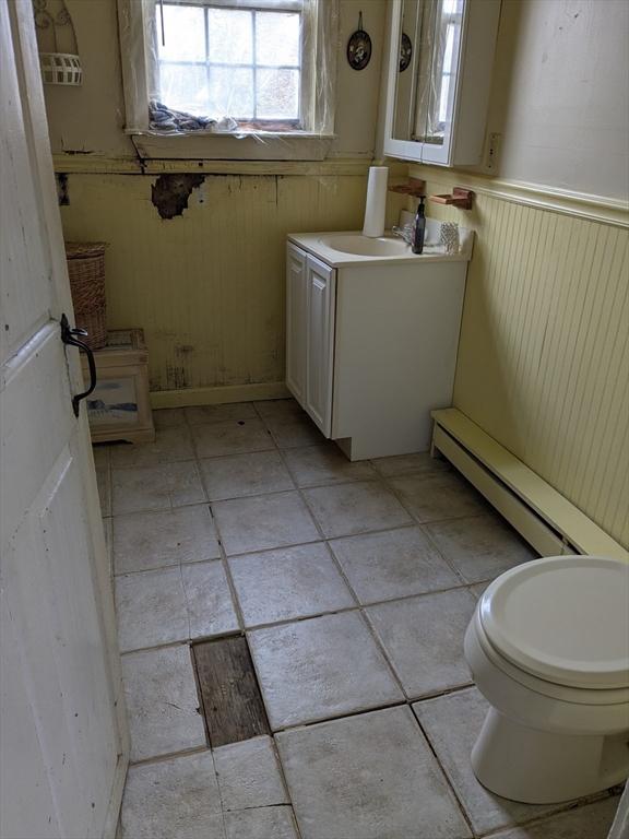 half bath with vanity, a wainscoted wall, a baseboard heating unit, toilet, and wooden walls
