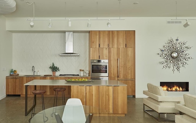 kitchen featuring brown cabinetry, concrete flooring, wall chimney range hood, a sink, and stainless steel appliances