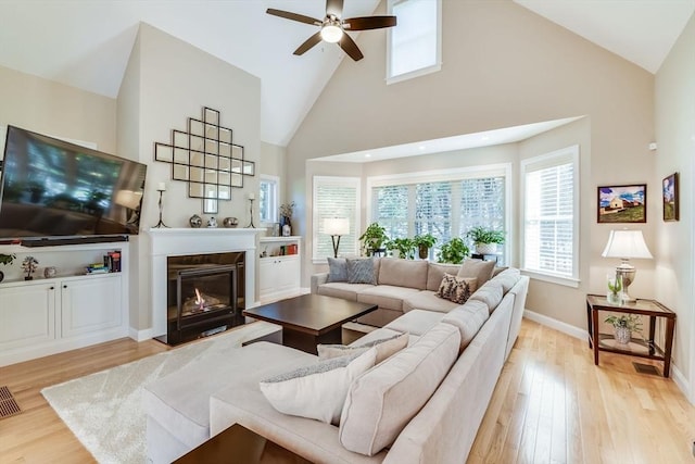 living area featuring baseboards, a fireplace with flush hearth, light wood-style flooring, high vaulted ceiling, and a ceiling fan