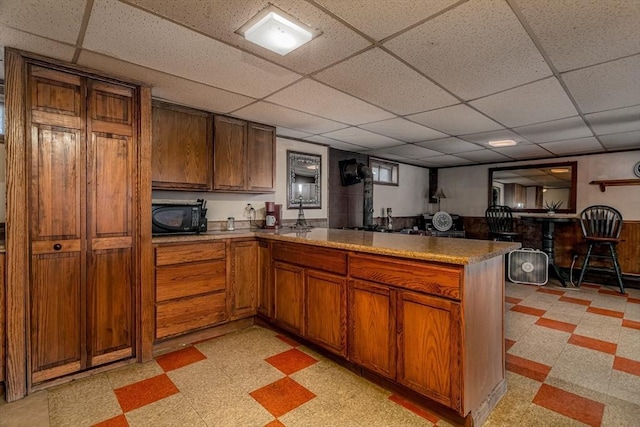 kitchen with a peninsula, black microwave, a drop ceiling, and light floors