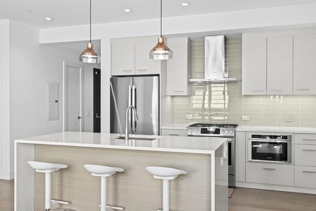 kitchen featuring appliances with stainless steel finishes, white cabinets, backsplash, wall chimney range hood, and a center island with sink