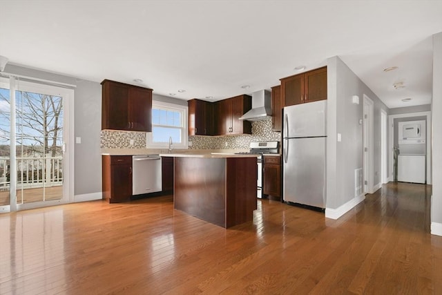 kitchen with stacked washer and dryer, a kitchen island, light countertops, appliances with stainless steel finishes, and wall chimney exhaust hood