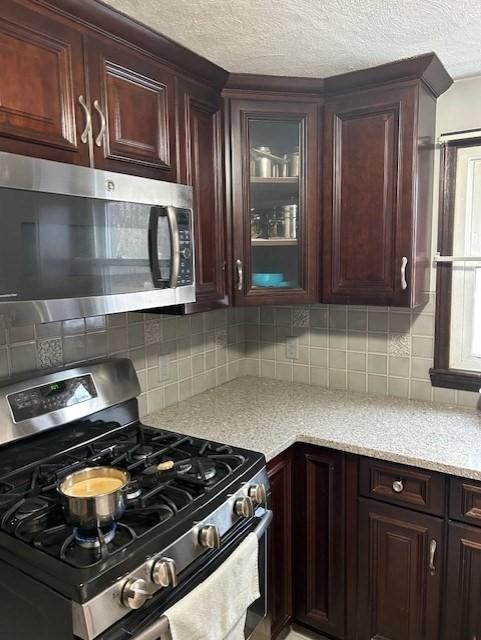 kitchen with light stone counters, decorative backsplash, appliances with stainless steel finishes, glass insert cabinets, and a textured ceiling