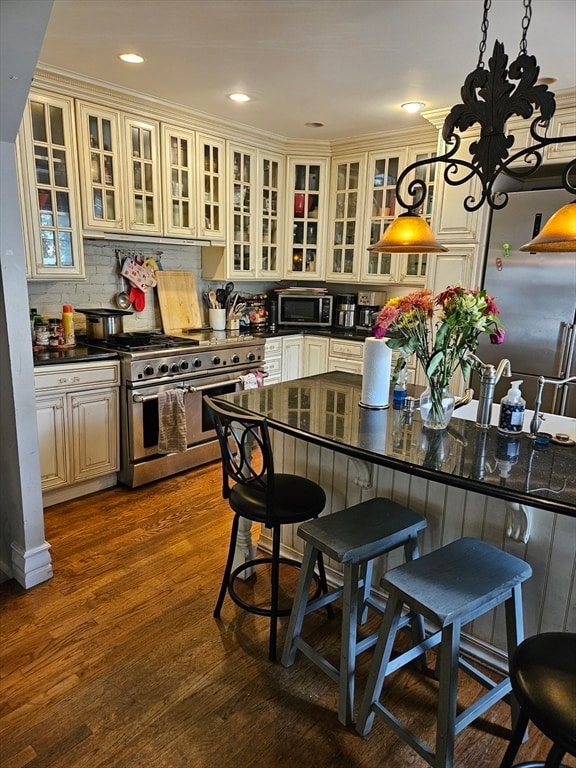 kitchen with cream cabinetry, ornamental molding, pendant lighting, appliances with stainless steel finishes, and dark hardwood / wood-style flooring