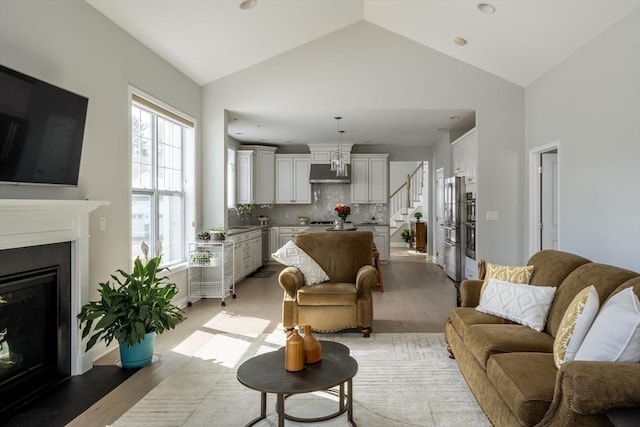 living room with a glass covered fireplace, high vaulted ceiling, light wood-type flooring, baseboards, and stairs