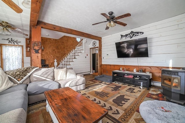 living room with wood walls, a wood stove, a baseboard radiator, beamed ceiling, and ceiling fan