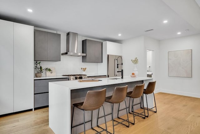 kitchen with wall chimney range hood, high end refrigerator, a kitchen island with sink, light wood finished floors, and recessed lighting