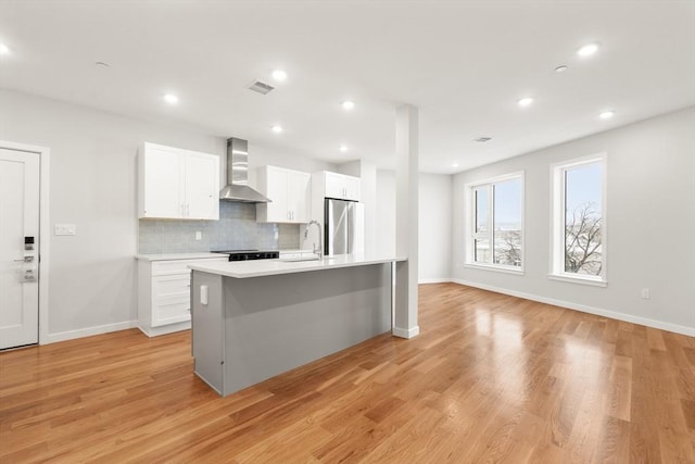 kitchen with a center island with sink, wall chimney exhaust hood, freestanding refrigerator, light countertops, and white cabinetry