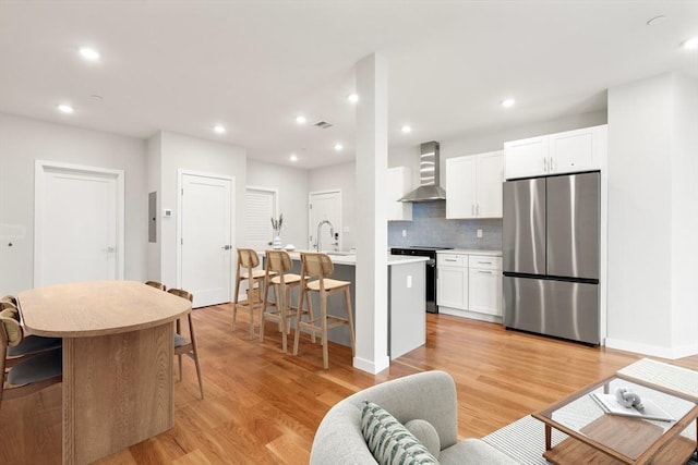 kitchen featuring electric range, freestanding refrigerator, a kitchen island with sink, white cabinets, and wall chimney range hood