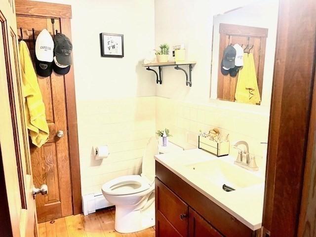 half bath featuring toilet, a baseboard heating unit, wood finished floors, vanity, and tile walls