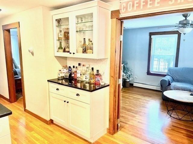 bar with a dry bar, baseboard heating, light wood-style floors, ceiling fan, and baseboards