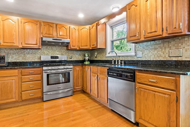 kitchen with stainless steel appliances, under cabinet range hood, light wood finished floors, dark stone counters, and a sink