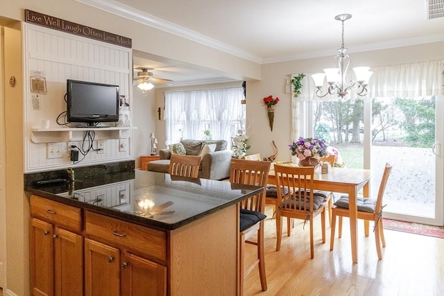 kitchen with brown cabinets, hanging light fixtures, light wood-style flooring, dark stone countertops, and ornamental molding