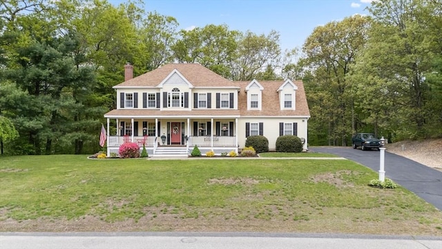 colonial inspired home with covered porch, a front lawn, a chimney, asphalt driveway, and view of wooded area